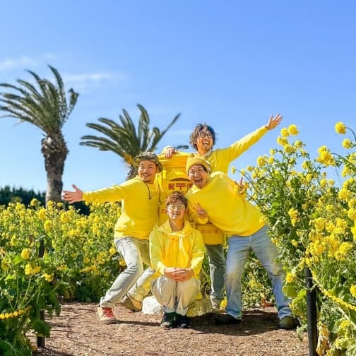 満開の菜の花畑と青空をバックに、お揃いの黄色いトップスを着てポーズをとるスタッフたち